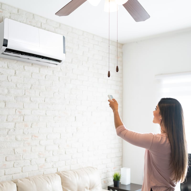 Femme souriante réglant un climatiseur mural blanc avec une télécommande. Un salon moderne avec mur en briques blanches et ventilateur de plafond.