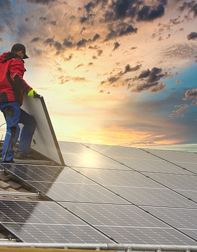 Un installateur pose un panneau solaire sur un toit. Le soleil couchant éclaire les nombreux panneaux solaires déjà installés.