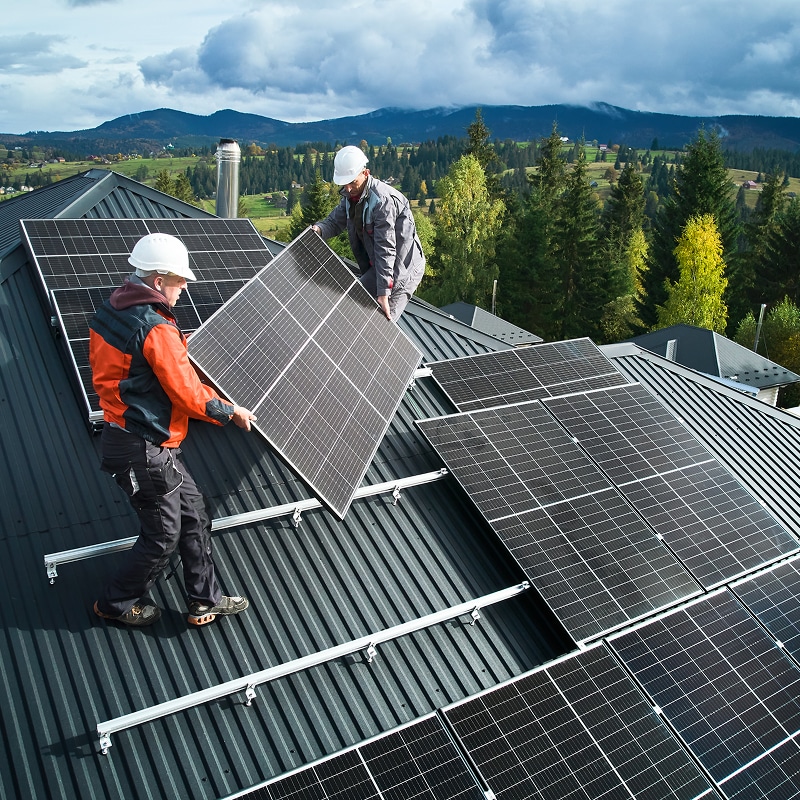 Deux techniciens installent des panneaux solaires sur un toit de maison. D'autres panneaux sont déjà posés, avec des montagnes en fond.